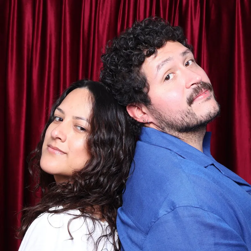 Dan and Alexis standing back to back in front of a red curtain, looking at the camera